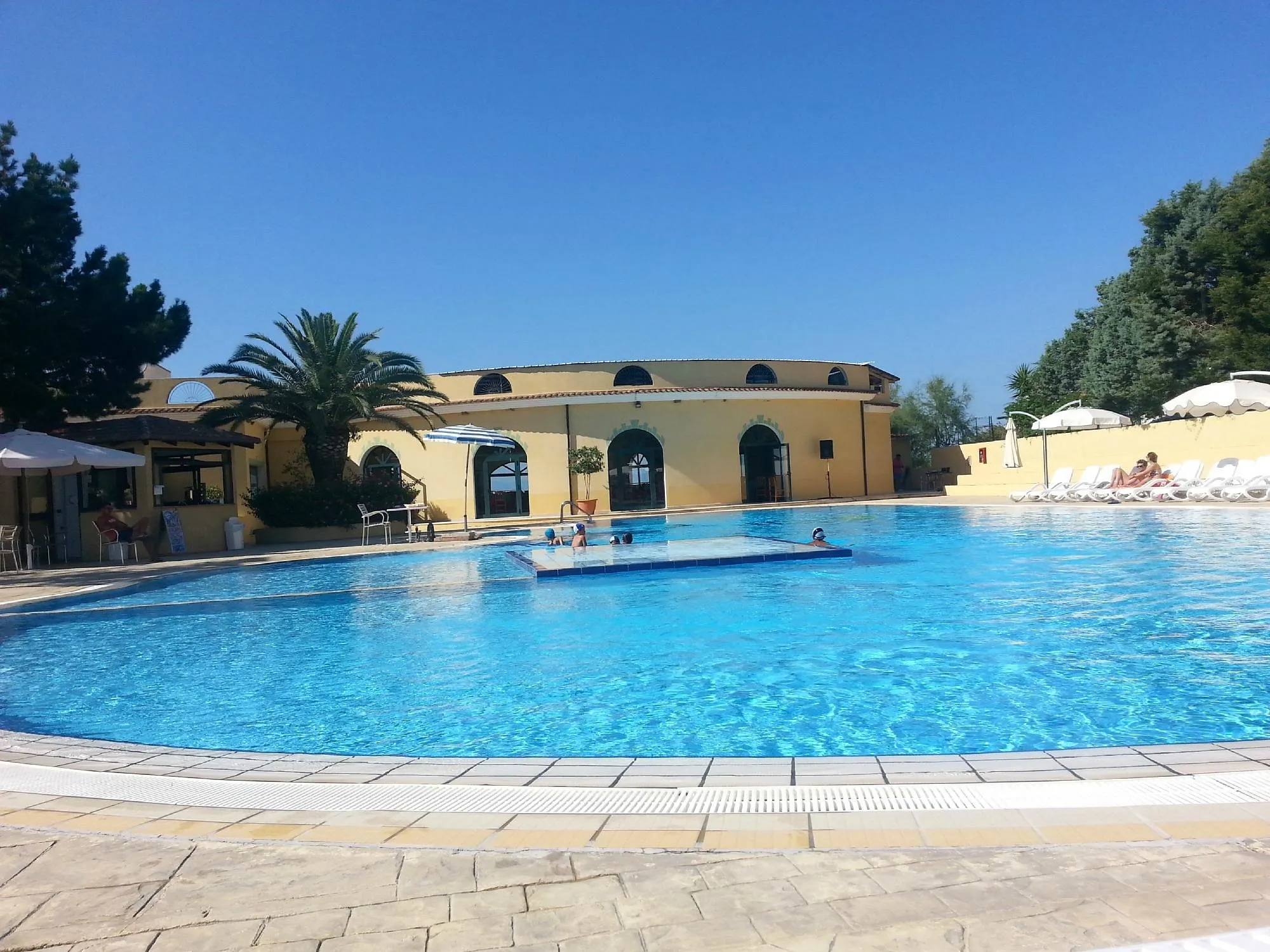 Piscina con bagnanti e edificio giallo del resort con palma e ombrelloni