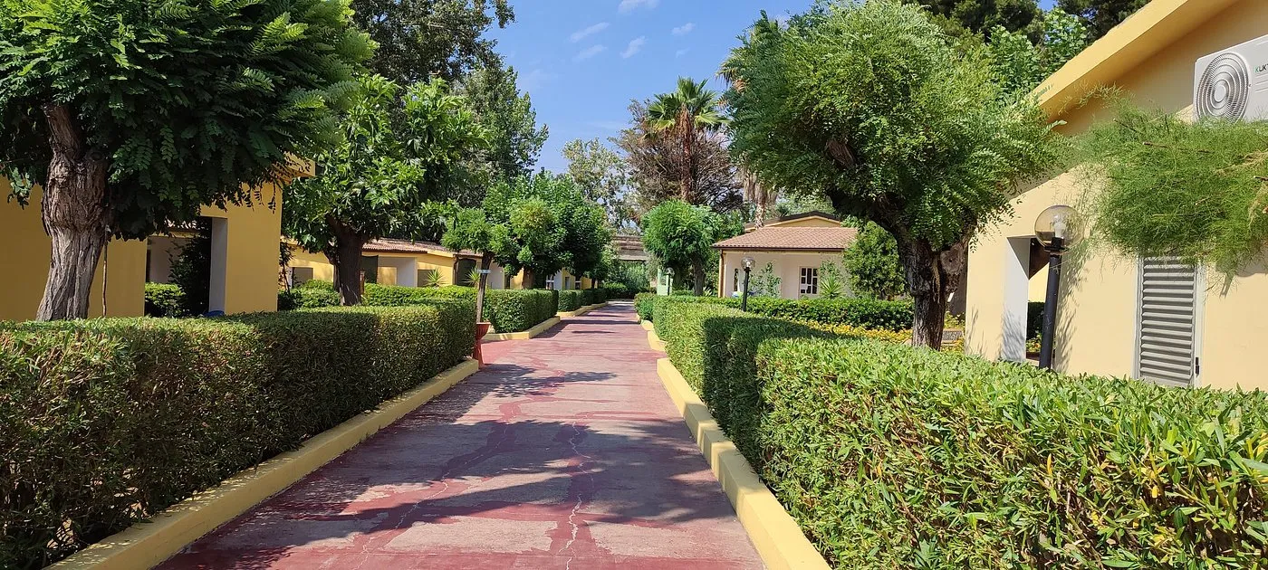 Vialetto rosso tra siepi curate, alberi e bungalow gialli del residence